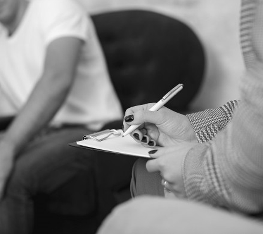 Une femme prend des notes en avant de l'image, et au second plan un homme se confie. L'image est en noir et blanc. Nous ne voyons que les torses et bras des personnages.