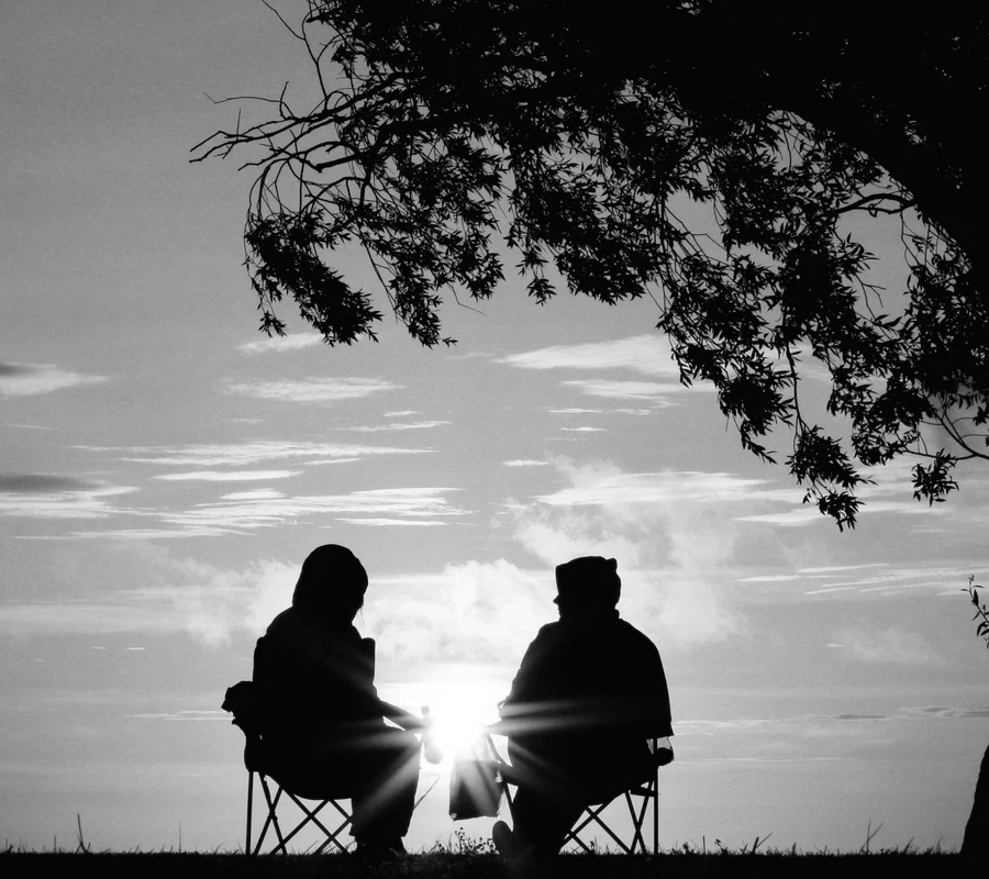 2 silhouettes de personnes discutent au pied d'un arbre sur des chaises de camping au lever du soleil, l'un semble se confier à l'autre. L'image est en noir et blanc.